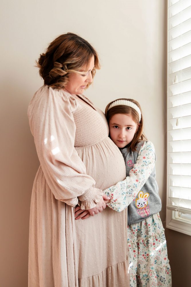 Cleveland maternity photography session of a pregnant mother with her young daughter, captured in soft natural window light by Allison Liffman Photography, highlighting a growing family and sibling connection in a warm, neutral-toned indoor setting