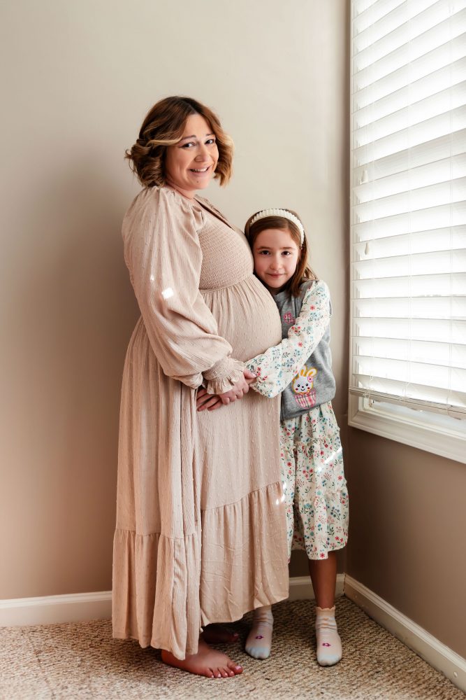 Cleveland maternity photography session of a pregnant mother with her young daughter, captured in soft natural window light by Allison Liffman Photography, highlighting a growing family and sibling connection in a warm, neutral-toned indoor setting