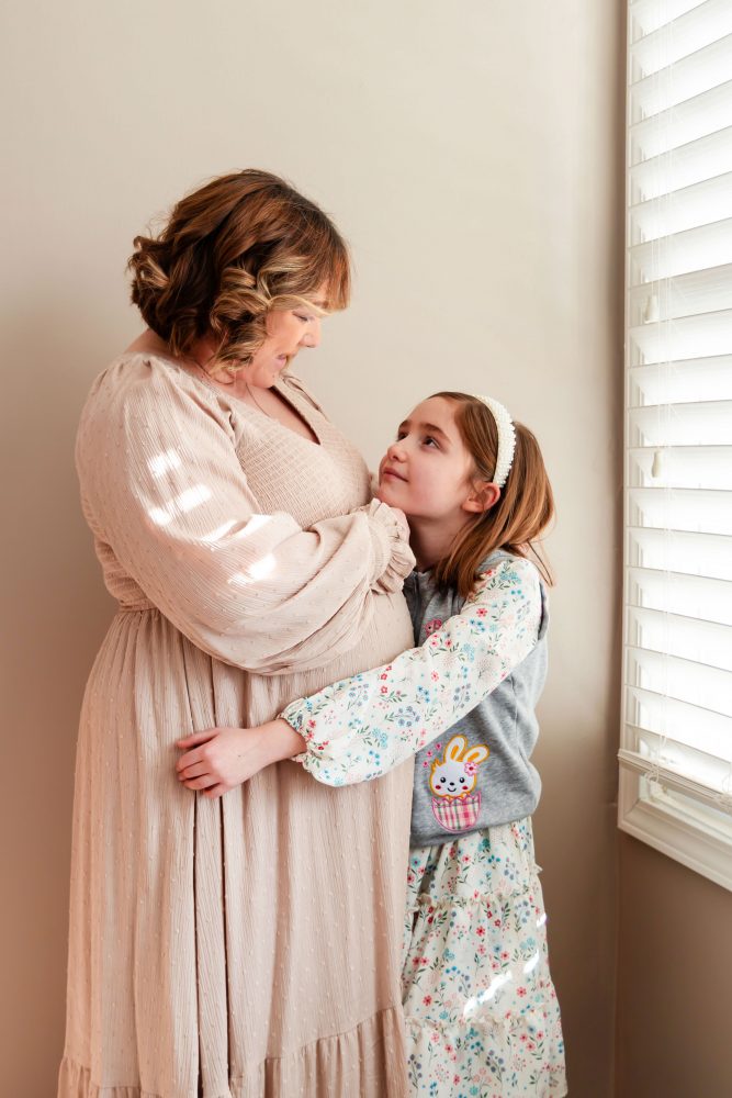 Cleveland maternity photography session of a pregnant mother with her young daughter, captured in soft natural window light by Allison Liffman Photography, highlighting a growing family and sibling connection in a warm, neutral-toned indoor setting