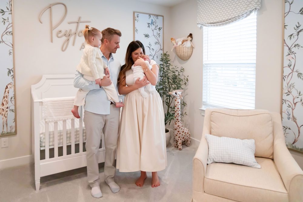 Family of four standing together in a neutral-toned nursery, parents holding their newborn son while big sister cuddles with dad during an in-home newborn photography session in Cleveland.