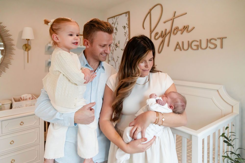 Family of four standing together in a neutral-toned nursery, parents holding their newborn son while big sister cuddles with dad during an in-home newborn photography session in Cleveland.