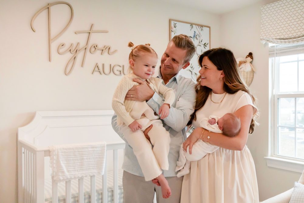 Family of four standing together in a neutral-toned nursery, parents holding their newborn son while big sister cuddles with dad during an in-home newborn photography session in Cleveland.
