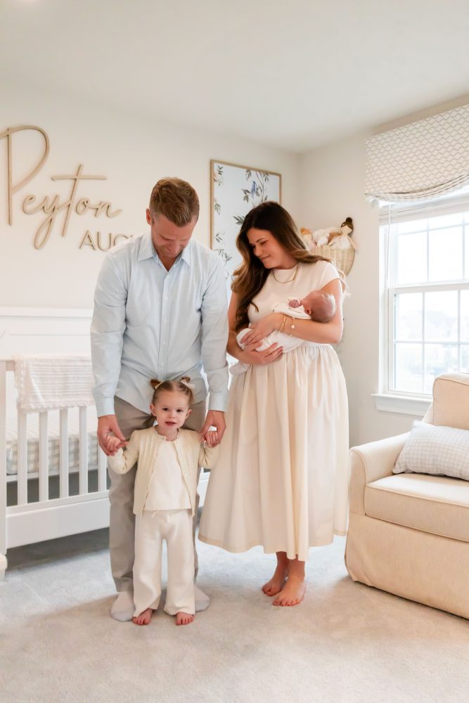 Family of four standing together in a neutral-toned nursery, parents holding their newborn son while big sister cuddles with dad during an in-home newborn photography session in Cleveland.