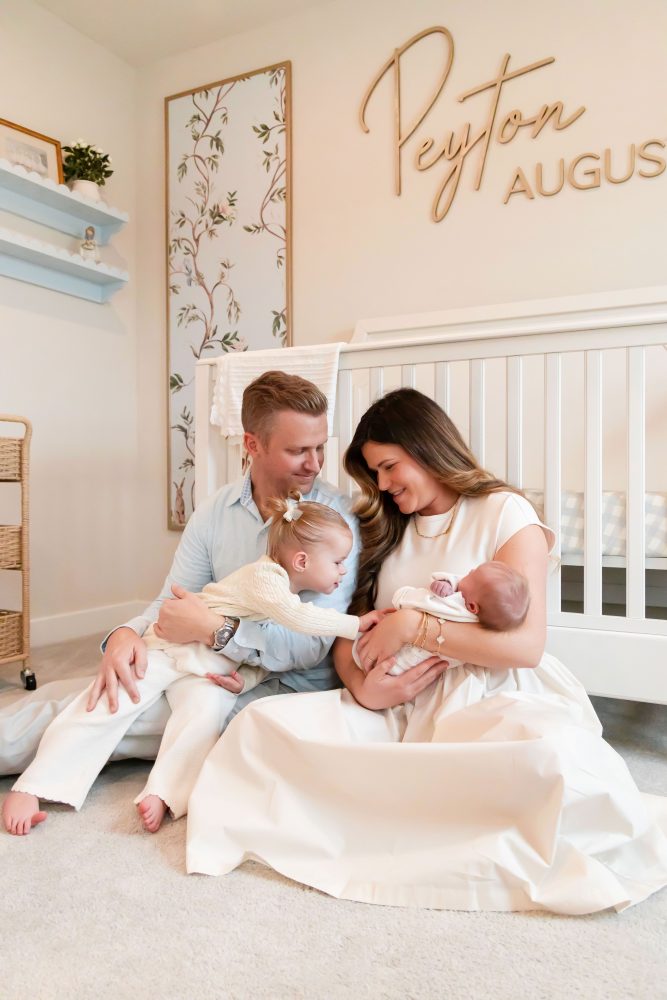 Family of four standing together in a neutral-toned nursery, parents holding their newborn son while big sister cuddles with dad during an in-home newborn photography session in Cleveland.