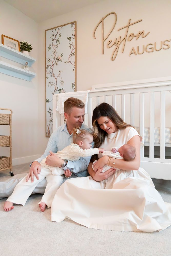 Family of four standing together in a neutral-toned nursery, parents holding their newborn son while big sister cuddles with dad during an in-home newborn photography session in Cleveland.