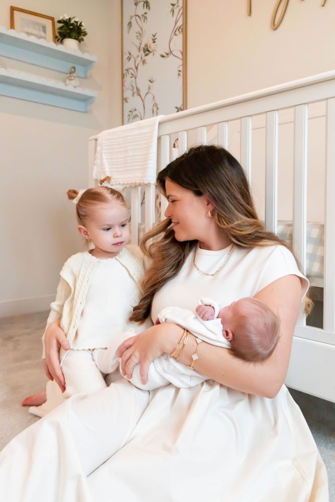 Parents sitting on the nursery floor with their newborn baby boy while big sister gently reaches toward her new sibling beside a white crib and custom nursery decor.