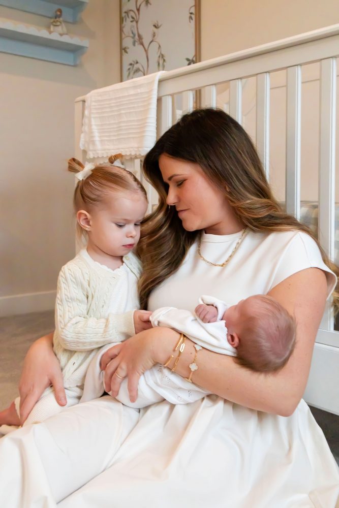 Parents sitting on the nursery floor with their newborn baby boy while big sister gently reaches toward her new sibling beside a white crib and custom nursery decor.