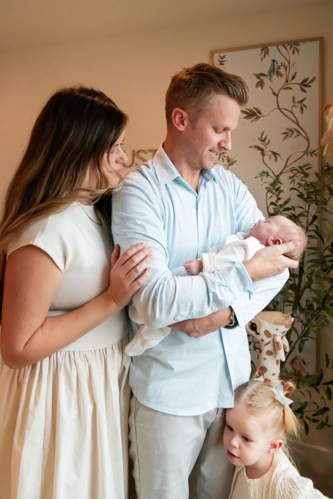 Family of four standing together in a neutral-toned nursery, parents holding their newborn son while big sister cuddles with dad during an in-home newborn photography session in Cleveland.