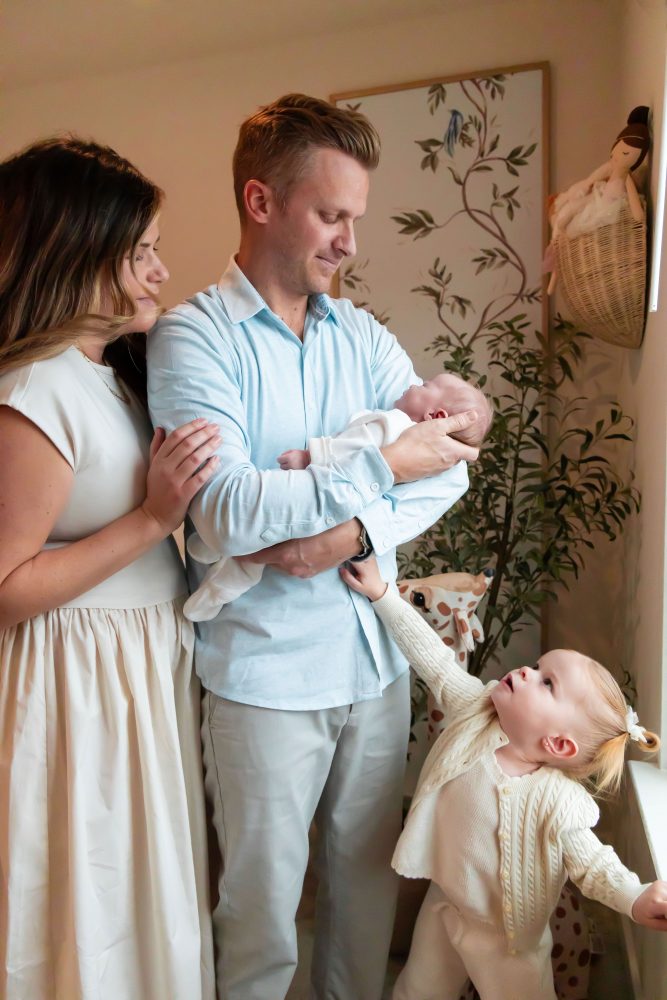 Family of four standing together in a neutral-toned nursery, parents holding their newborn son while big sister cuddles with dad during an in-home newborn photography session in Cleveland.