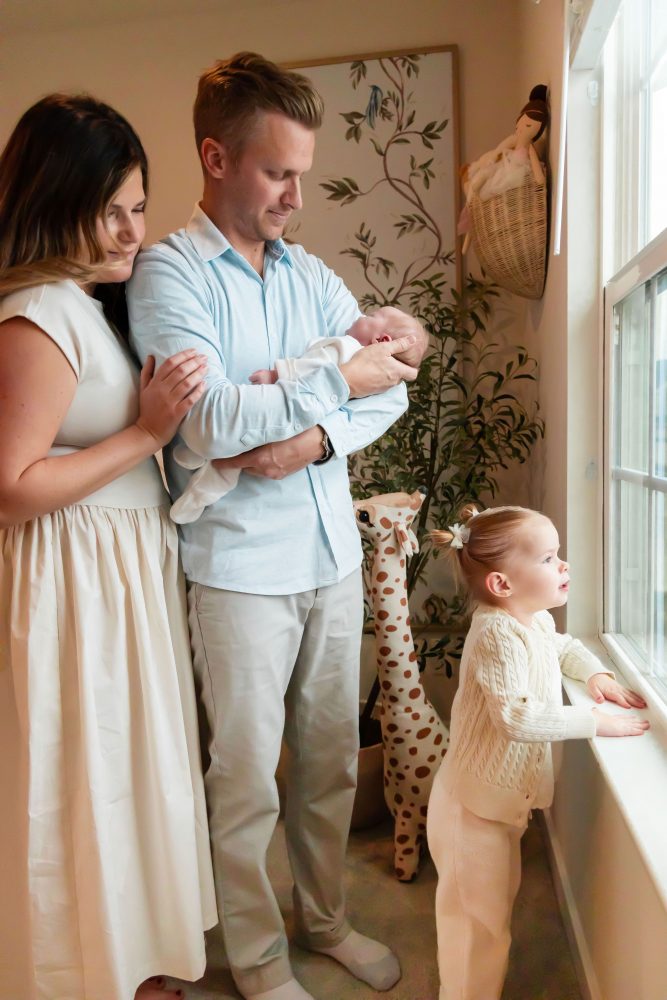Family of four standing together in a neutral-toned nursery, parents holding their newborn son while big sister cuddles with dad during an in-home newborn photography session in Cleveland.