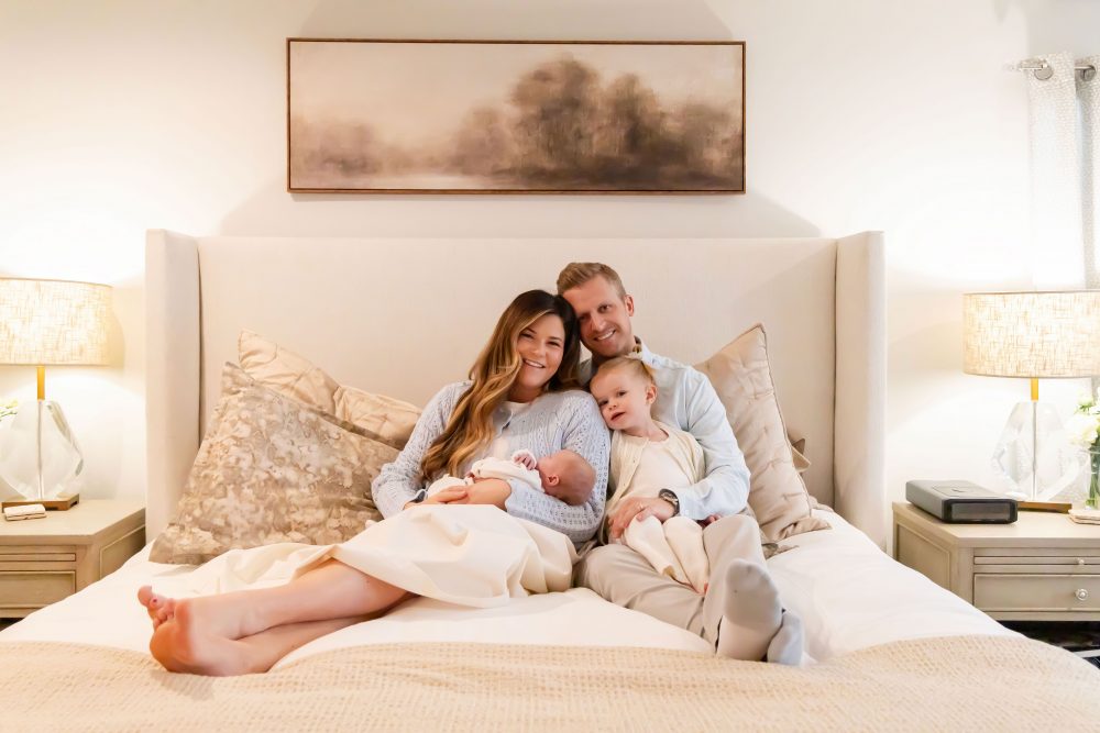 Lifestyle family portrait on a bed featuring newborn baby in the foreground with parents and toddler smiling softly in the background during a cozy in-home newborn session.