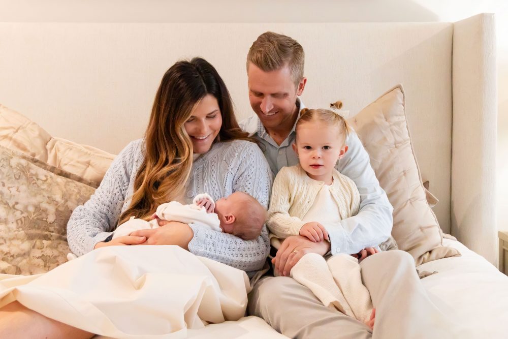 Lifestyle family portrait on a bed featuring newborn baby in the foreground with parents and toddler smiling softly in the background during a cozy in-home newborn session.