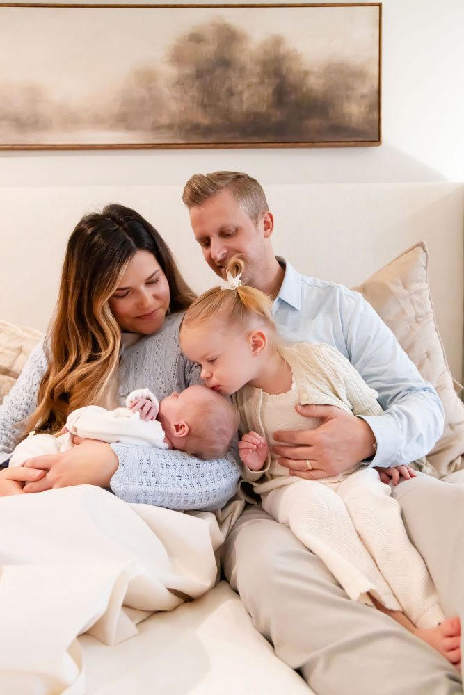 Lifestyle family portrait on a bed featuring newborn baby in the foreground with parents and toddler smiling softly in the background during a cozy in-home newborn session.