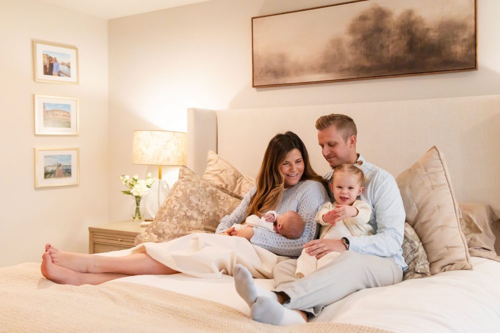 Lifestyle family portrait on a bed featuring newborn baby in the foreground with parents and toddler smiling softly in the background during a cozy in-home newborn session.