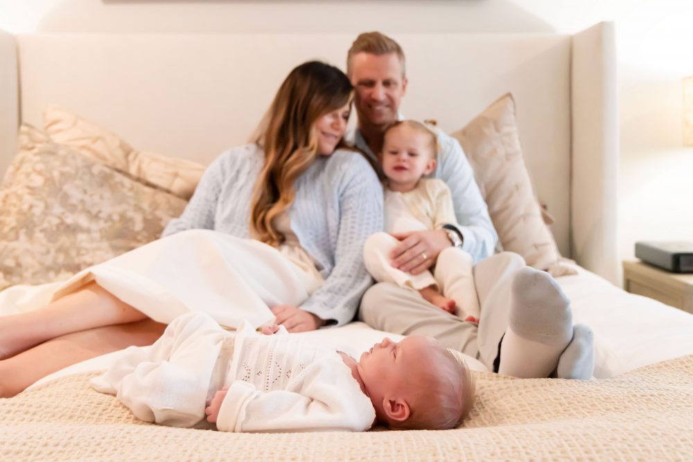 Lifestyle family portrait on a bed featuring newborn baby in the foreground with parents and toddler smiling softly in the background during a cozy in-home newborn session.