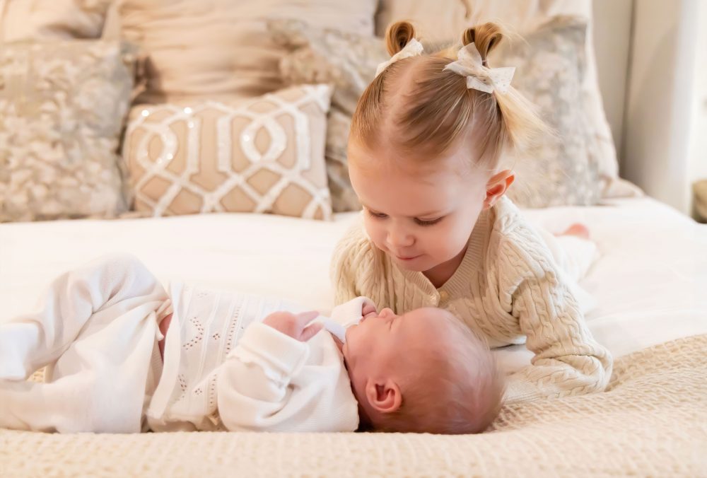 Lifestyle family portrait on a bed featuring newborn baby in the foreground with toddler smiling softly in the background during a cozy in-home newborn session.