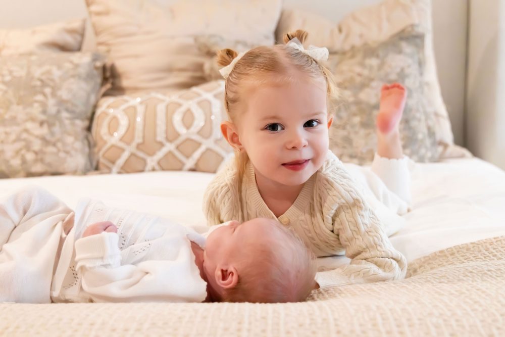 Lifestyle family portrait on a bed featuring newborn baby in the foreground with toddler smiling softly in the background during a cozy in-home newborn session.