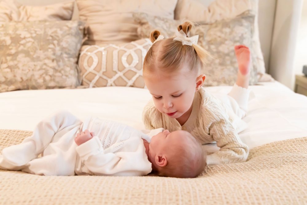 Lifestyle family portrait on a bed featuring newborn baby in the foreground with toddler smiling softly in the background during a cozy in-home newborn session.
