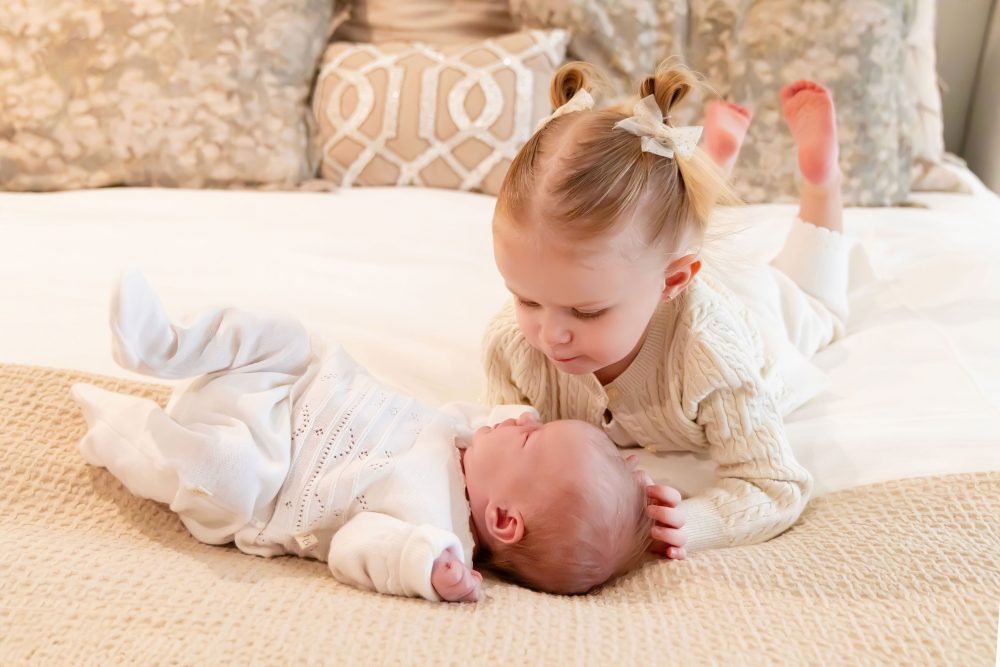 Lifestyle family portrait on a bed featuring newborn baby in the foreground with toddler smiling softly in the background during a cozy in-home newborn session.
