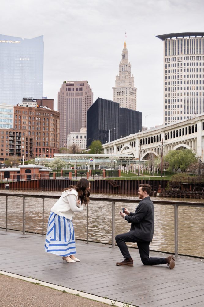 Couple celebrating a surprise proposal on a downtown Cleveland waterfront boardwalk with the Cleveland skyline in the background