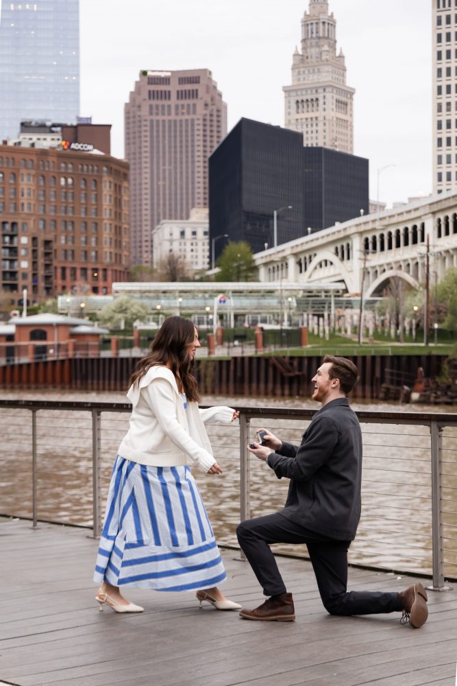 Couple celebrating a surprise proposal on a downtown Cleveland waterfront boardwalk with the Cleveland skyline in the background