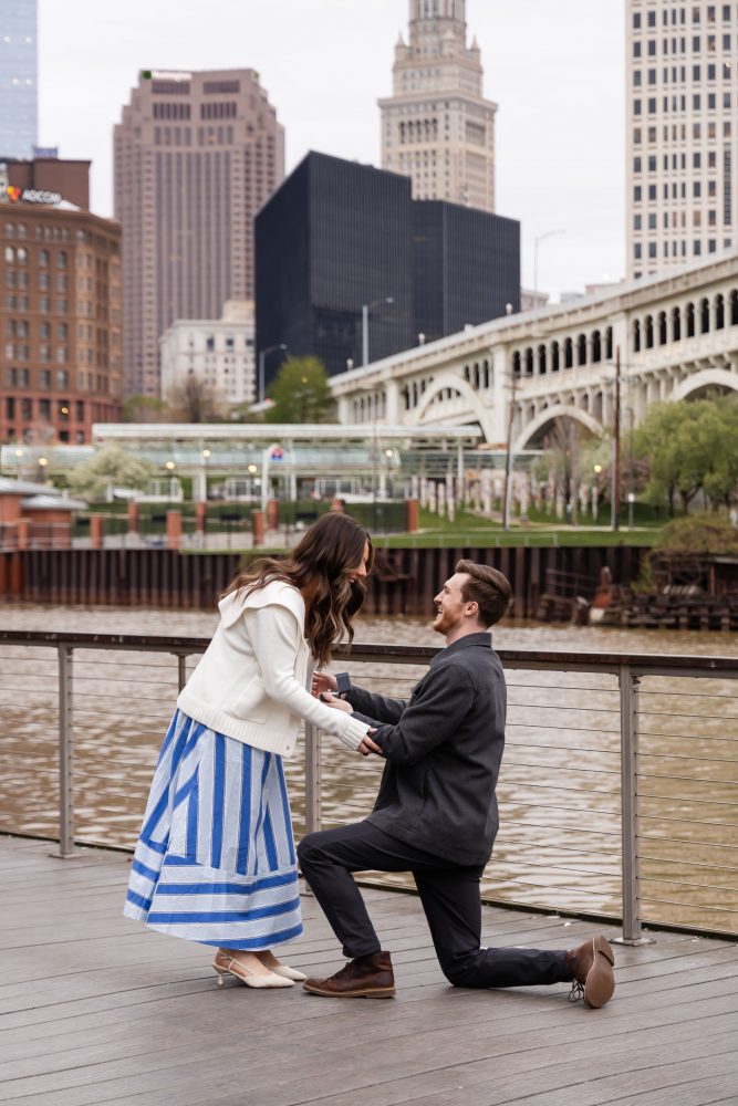 Couple celebrating a surprise proposal on a downtown Cleveland waterfront boardwalk with the Cleveland skyline in the background