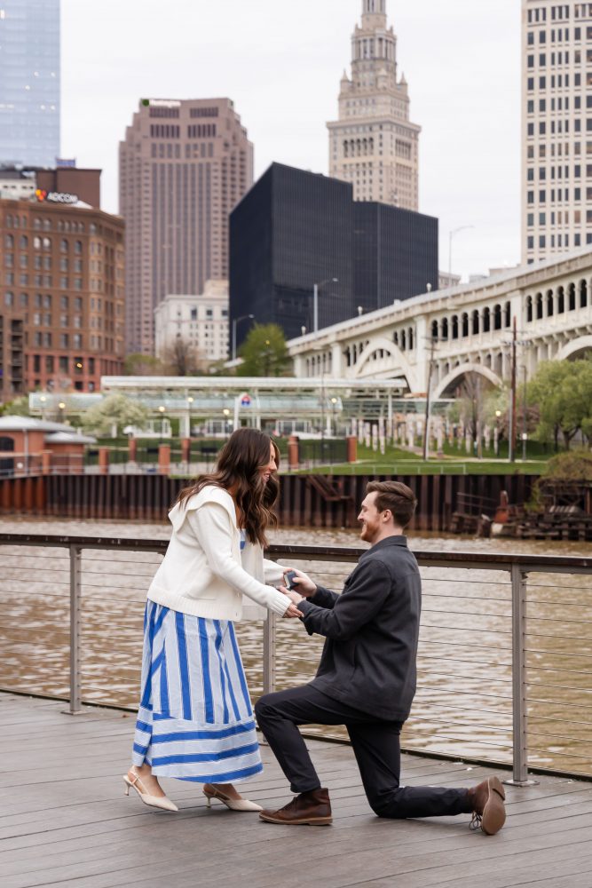 Couple celebrating a surprise proposal on a downtown Cleveland waterfront boardwalk with the Cleveland skyline in the background