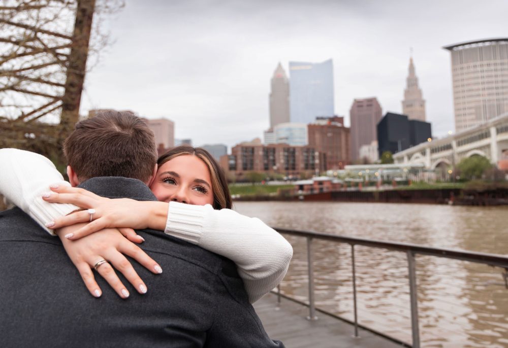Couple celebrating a surprise proposal on a downtown Cleveland waterfront boardwalk with the Cleveland skyline in the background