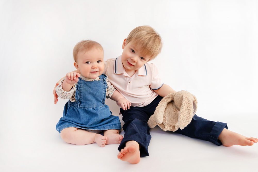 Older brother sitting with baby sister in a studio, smiling and cuddling, neutral white background family portrait