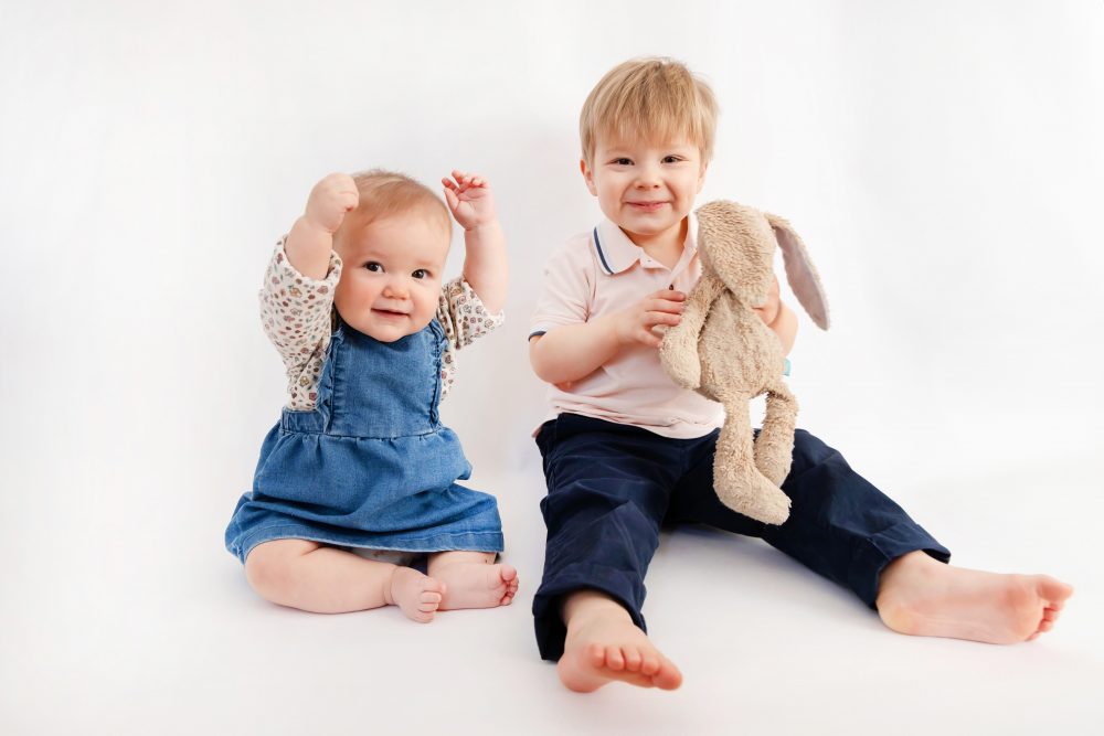 Older brother sitting with baby sister in a studio, smiling and cuddling, neutral white background family portrait