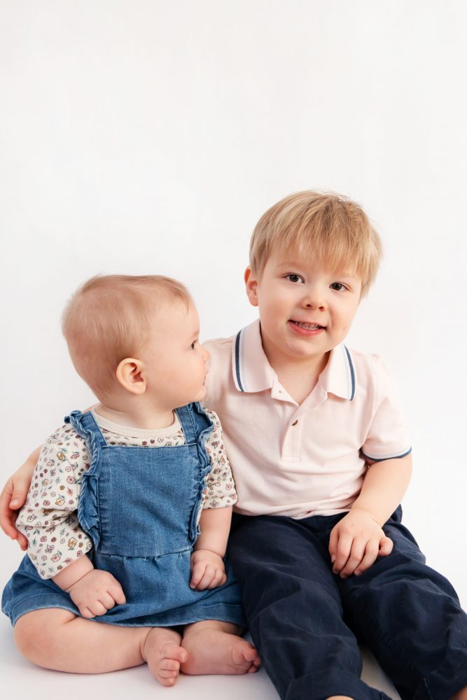 Older brother sitting with baby sister in a studio, smiling and cuddling, neutral white background family portrait