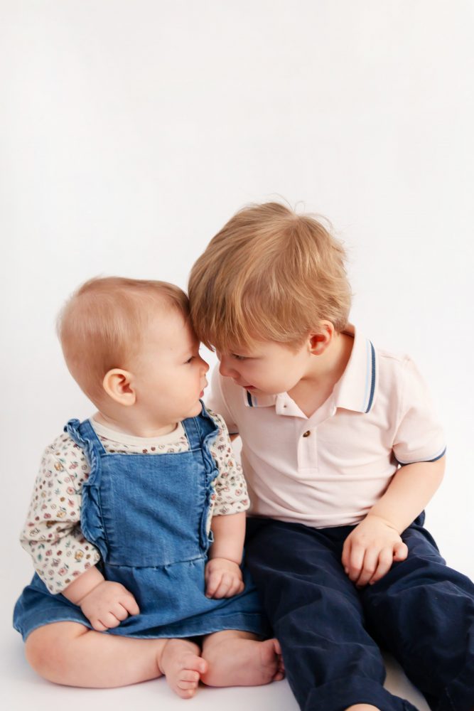 Older brother sitting with baby sister in a studio, smiling and cuddling, neutral white background family portrait