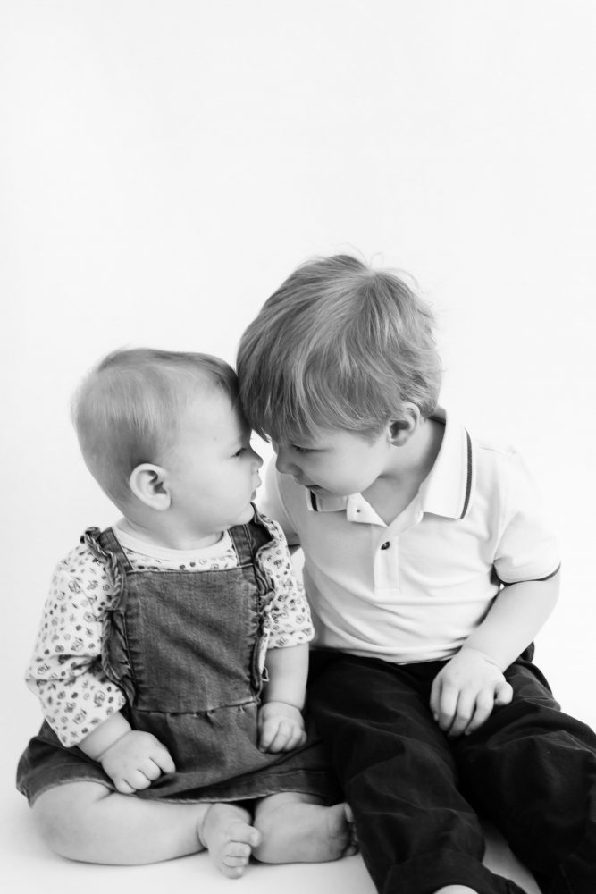 Older brother sitting with baby sister in a studio, smiling and cuddling, neutral white background family portrait