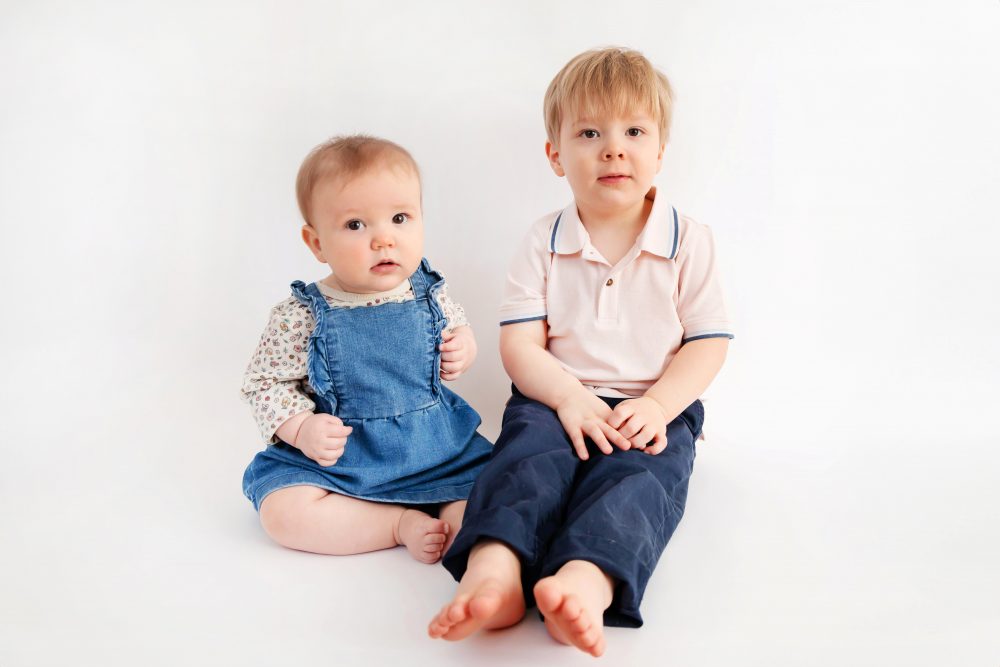 Older brother sitting with baby sister in a studio, smiling and cuddling, neutral white background family portrait