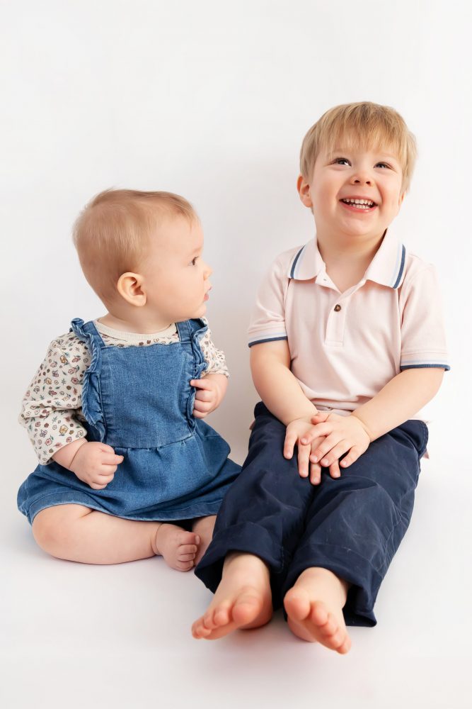 Older brother sitting with baby sister in a studio, smiling and cuddling, neutral white background family portrait