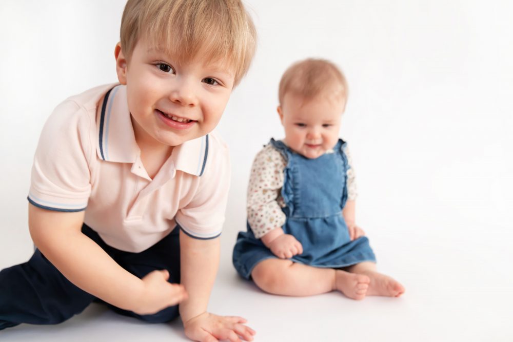 Older brother sitting with baby sister in a studio, smiling and cuddling, neutral white background family portrait