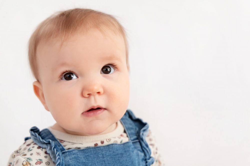 Baby girl in a blue dress crawling and smiling during a studio milestone session on a white backdrop