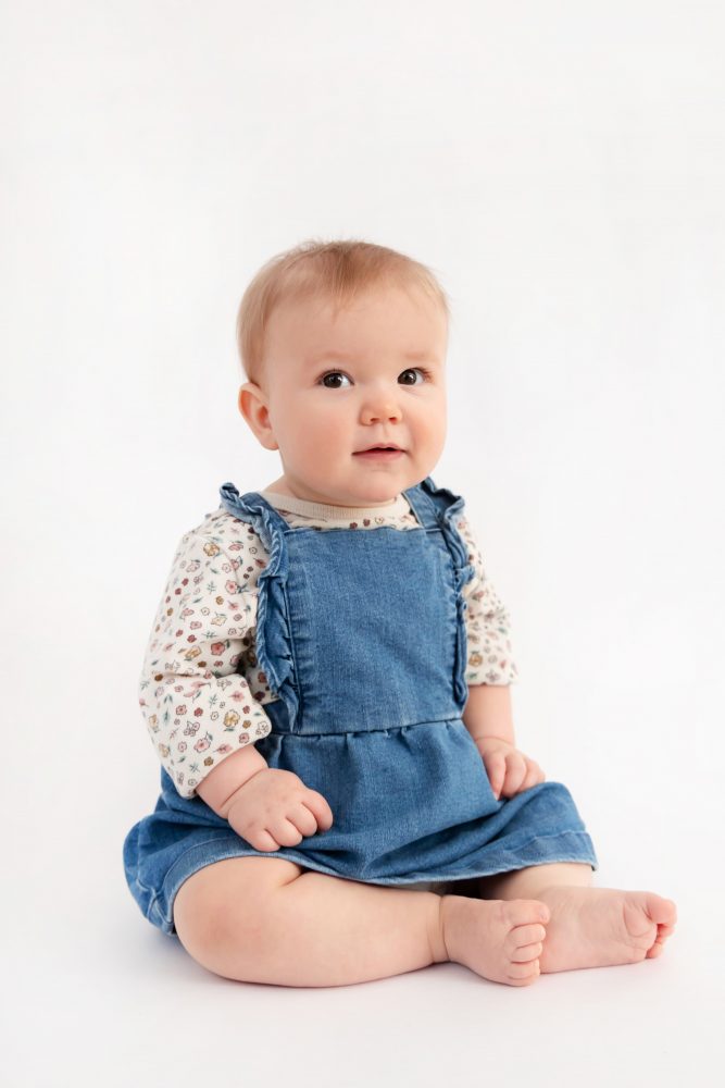 Baby girl in a blue dress crawling and smiling during a studio milestone session on a white backdrop