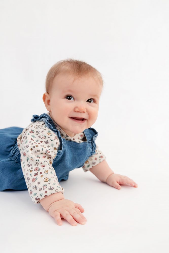 Baby girl in a blue dress crawling and smiling during a studio milestone session on a white backdrop