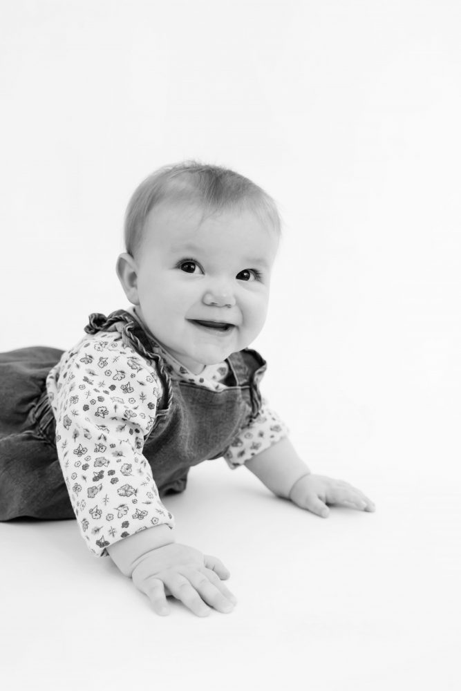 Baby girl in a blue dress crawling and smiling during a studio milestone session on a white backdrop
