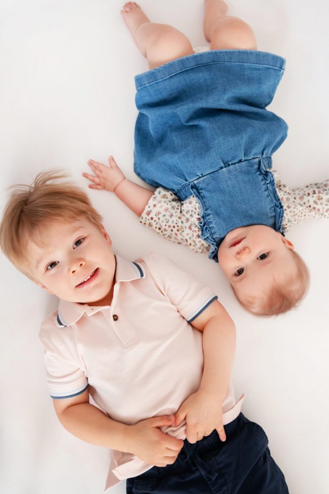Older brother sitting with baby sister in a studio, smiling and cuddling, neutral white background family portrait