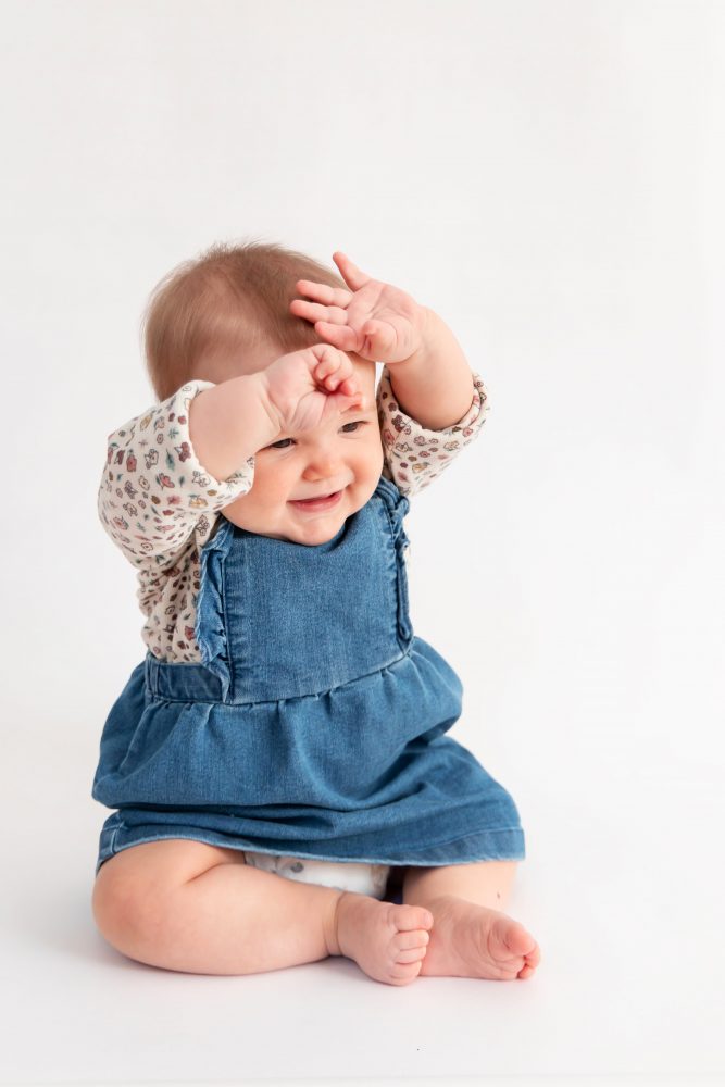 Baby girl in a blue dress crawling and smiling during a studio milestone session on a white backdrop