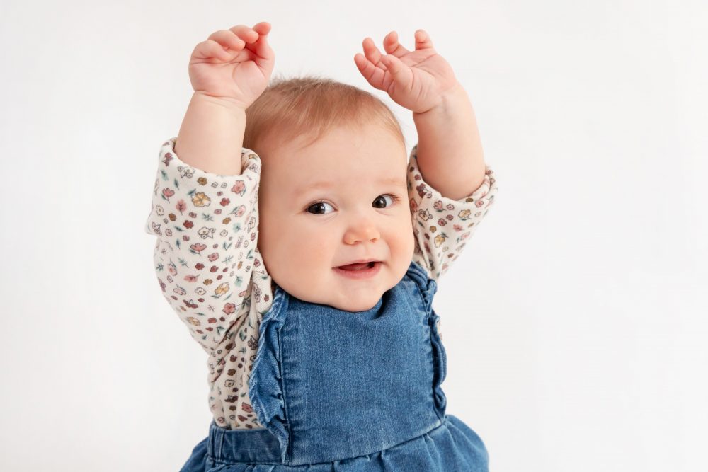 Baby girl in a blue dress crawling and smiling during a studio milestone session on a white backdrop