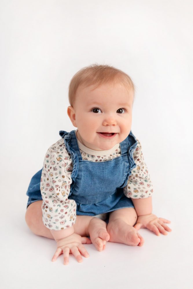 Baby girl in a blue dress crawling and smiling during a studio milestone session on a white backdrop