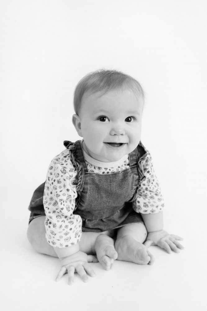 Baby girl in a blue dress crawling and smiling during a studio milestone session on a white backdrop