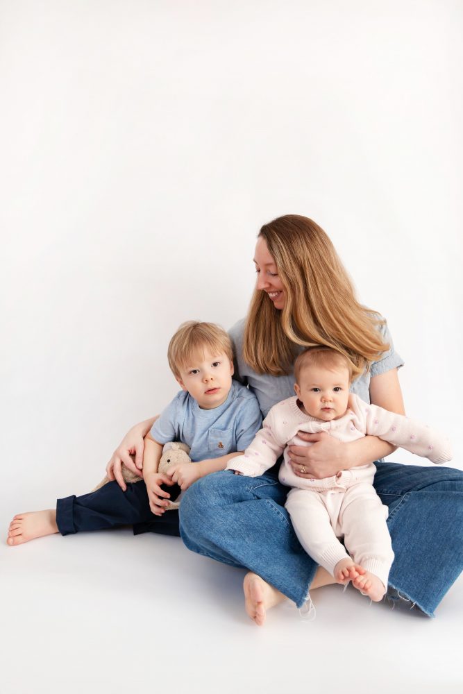 Older brother sitting with baby sister in a studio, smiling and cuddling, neutral white background family portrait