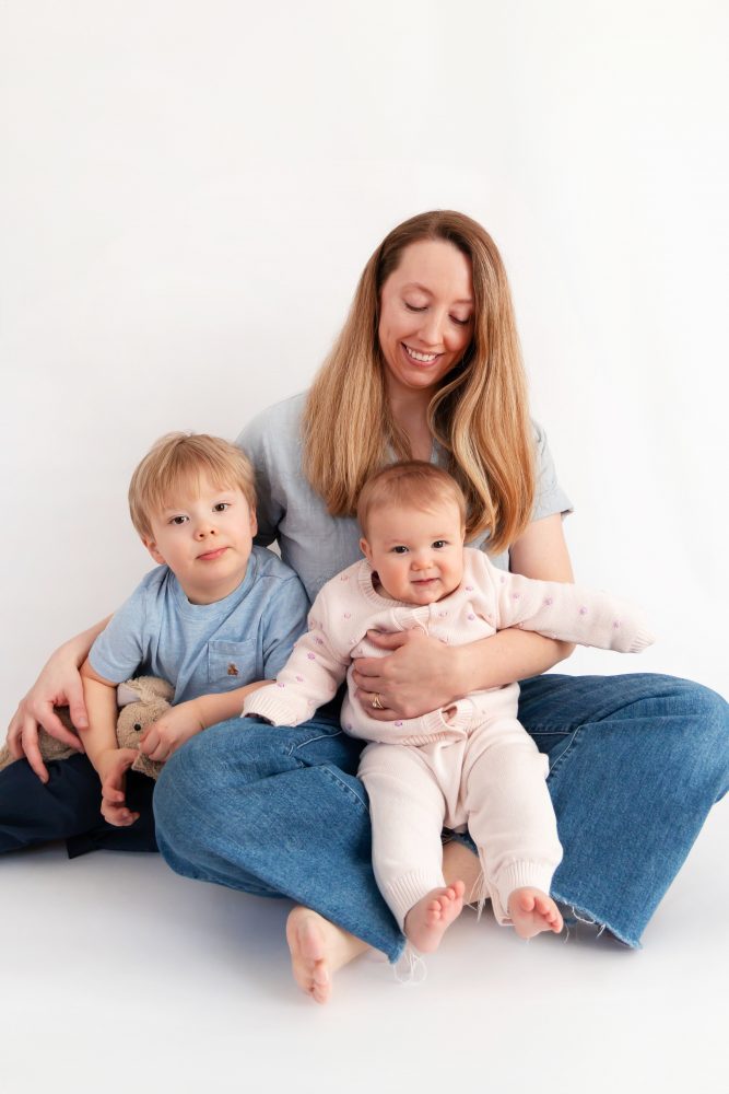 Older brother sitting with baby sister in a studio, smiling and cuddling, neutral white background family portrait