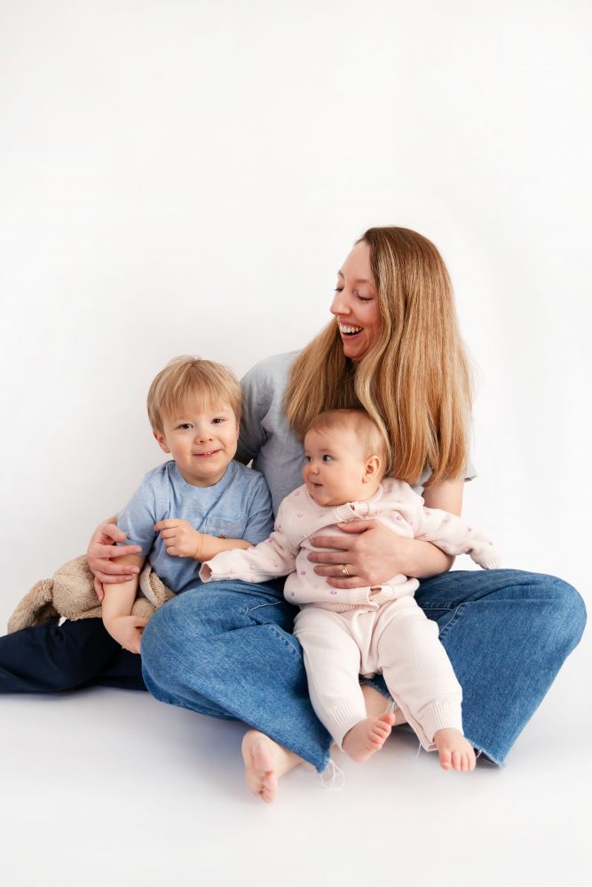 Older brother sitting with baby sister in a studio, smiling and cuddling, neutral white background family portrait
