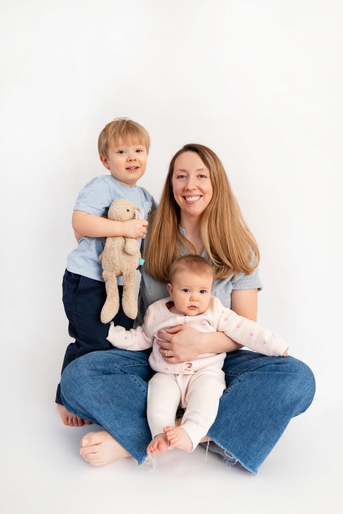 Older brother sitting with baby sister in a studio, smiling and cuddling, neutral white background family portrait
