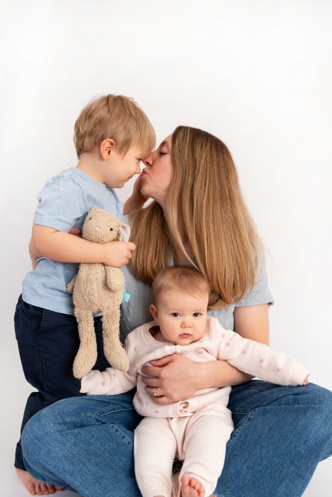Older brother sitting with baby sister in a studio, smiling and cuddling, neutral white background family portrait